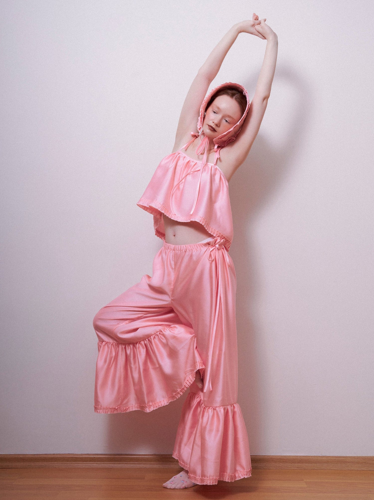 Person wearing a pink outfit with ruffled pants and top, standing against a plain wall.