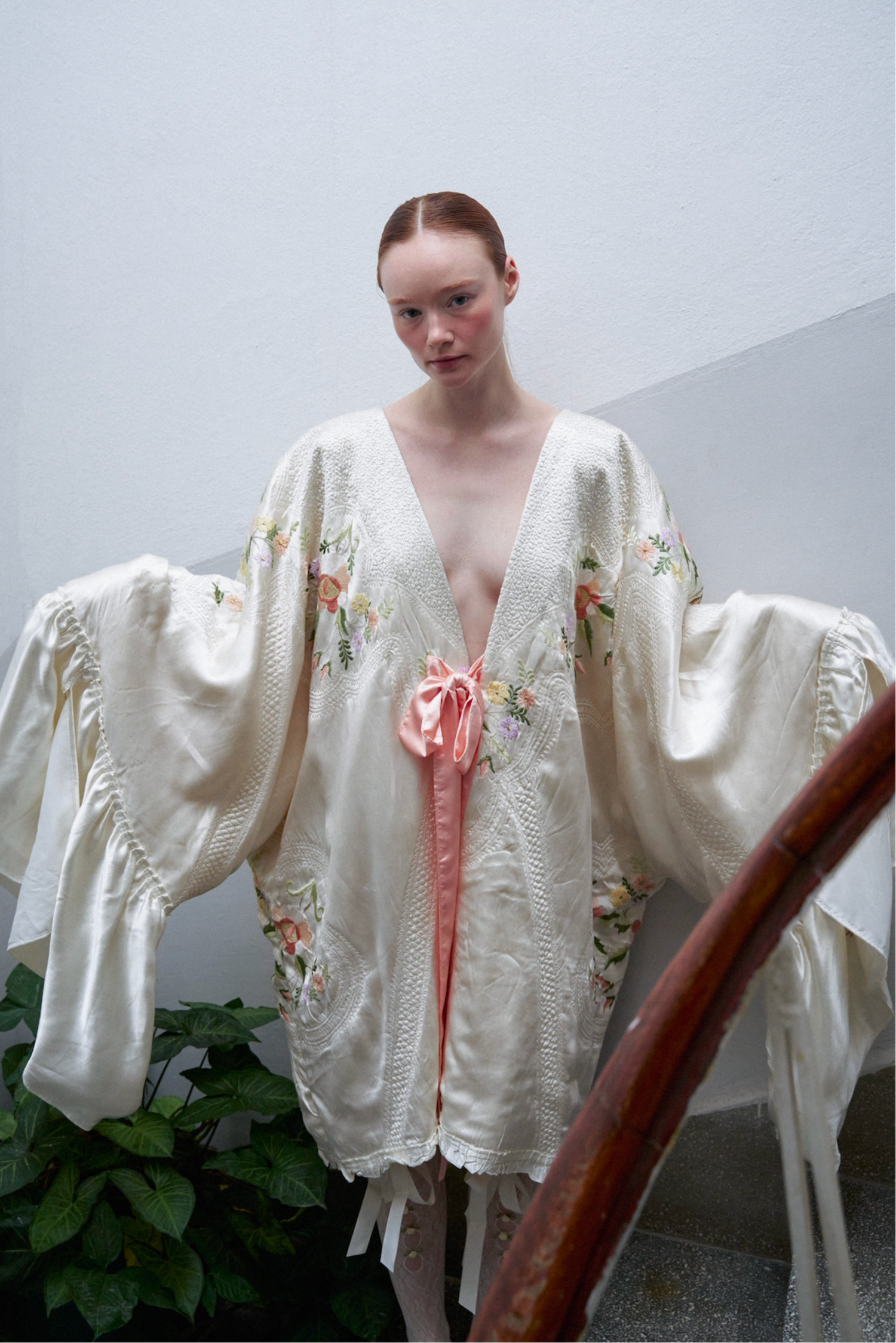 Woman wearing a white embroidered robe with a pink ribbon against a neutral background